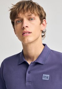 Lila polo skjorta med en texturerad tyg, treknapps knäppning och en broderad "POLO CLUB" patch på vänster bröst. Släta krage.