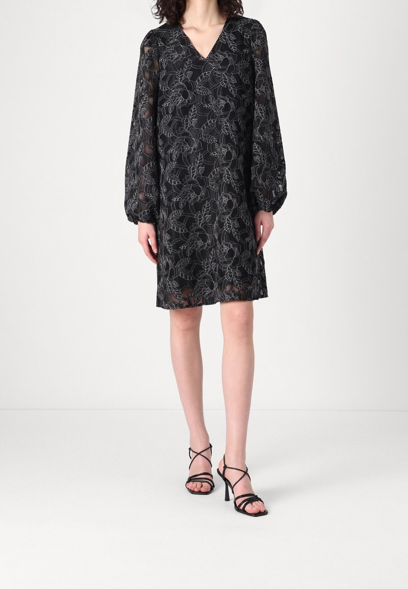 Femme portant une robe noire en dentelle florale à manches longues avec un col en V, associée à des sandales noires à talons hauts à brides, debout sur un sol blanc.