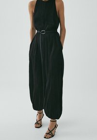 Musta haalari, jossa on niskalenkki, vyötäisellä kapea vyö, jossa on pyöreä solki, ja kapenevat housut. Yhdistetty mustien vyölenkkisten sandaalien kanssa.