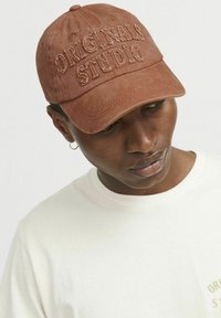 Brun bomulds kasket med broderet "ORIGINALS STUDIO" tekst. Har en buet skygge og justerbar rem. Bæres af en model iført en lys t-shirt.