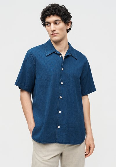 Άνδρας με σγουρά μαλλιά, ντυμένος με μπλε κοντομάνικο πουκάμισο με κουμπιά και ανοιχτό μπεζ παντελόνια, κοιτάζει στο πλάι με τα χέρια στις τσέπες.