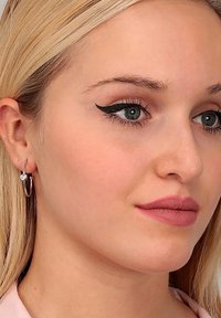 Orecchini a cerchio in argento con charm a forma di cuore, pelle liscia, eyeliner definito, labbra rosa chiaro, capelli biondi, sfondo neutro.