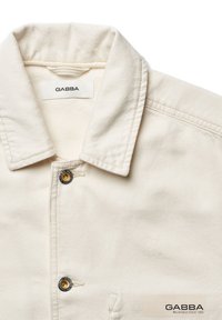 Beige denimjakke med spids krave, frontlukning med knapper og guldfarvede detaljer, samt et GABBA mærke ved halsen og nederste højre hjørne.