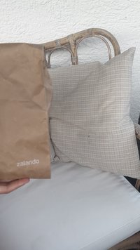 Rudas popierinis maišelis su užrašu "zalando" baltu spalva, laikomas priešais šachmatų rašto pagalvę, ant baltos pagalvės pintame krėsle.