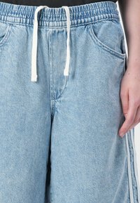 Joggers in denim leggero blu con cintura elastica e lacci bianchi. Presentano tasche laterali e strisce bianche ai lati. Texture liscia.