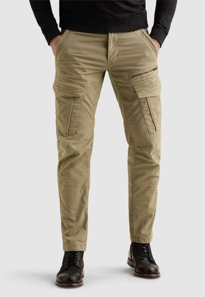 Homme portant un pantalon cargo kaki avec plusieurs poches et des bottines en cuir noir à lacets sur un fond uni.