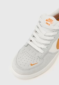 Sapatilha cinza em camurça com atacadores brancos, caixa de dedo perfurada, detalhe do logo Nike em laranja e sola branca texturizada.