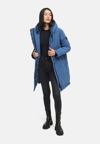 Manteau long bleu en duvet avec capuche, doté d'une doublure en fourrure, fermeture éclair et boutons-pression, associé à un jean skinny noir et des bottes noires.