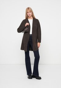 Cárdigan de punto marrón con abertura frontal, mangas largas, combinado con jeans acampanados oscuros y botas negras, sosteniendo un bolso pequeño. Camiseta blanca debajo.