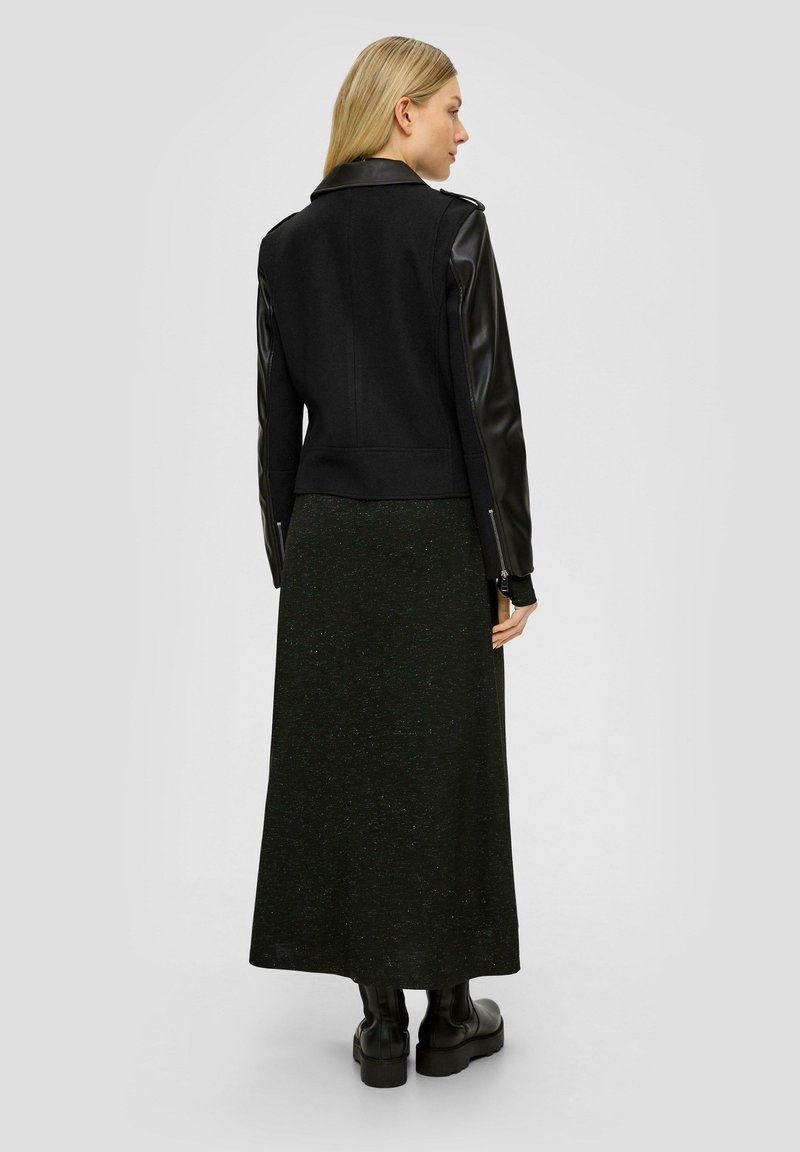 Schwarze Jacke mit einem Wollkörper und Lederärmeln, getragen über einem langen schwarzen Rock mit subtiler glänzender Oberfläche, kombiniert mit schwarzen Knöchelstiefeln.