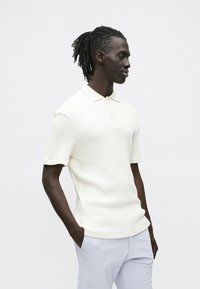 Polo bege creme com tecido canelado e mangas curtas, apresentando uma gola clássica e uma bainha reta. Combinada com calças de cor clara.