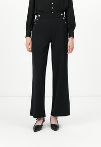 Pantalons larges noirs avec des détails en boutons dorés à la taille haute, associés à des chaussures à talons aiguilles noires et à un blouse noir à manches longues.