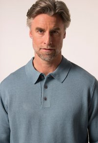 Jasnoniebieska dziergana koszulka polo z kołnierzykiem, trzema ciemnymi guzikami i teksturowanym wykończeniem. Materiał wydaje się miękki i wygodny.