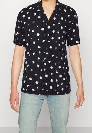 Homme portant une chemise noire à manches courtes avec des boutons et des pois blancs irréguliers, ainsi qu'un jean bleu clair, debout devant un fond uni.