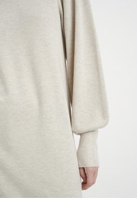 Robe beige à manches longues avec une texture douce, présentant une coupe décontractée et des manches bouffantes froncées. Les poignets côtelés soulignent le design.