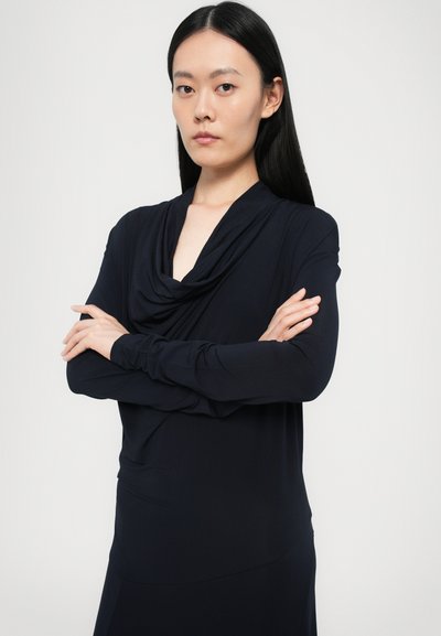 Femme aux longs cheveux noirs portant un haut noir à manches longues, debout les bras croisés devant un fond clair uni.