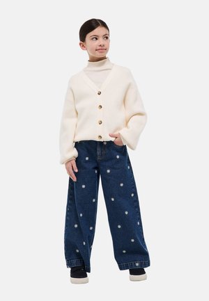 Jeune fille debout, main dans la poche, portant un cardigan crème boutonné, un col roulé blanc, un jean large orné d'imprimés de fleurs blanches, et des chaussures noires.