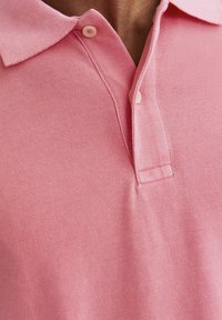 Polo majica v svetlo roza barvi z teksturirano tkanino, z zapenjanjem na tri gumbe in klasičnim ovratnikom. Njegov material je mehak in zračen.