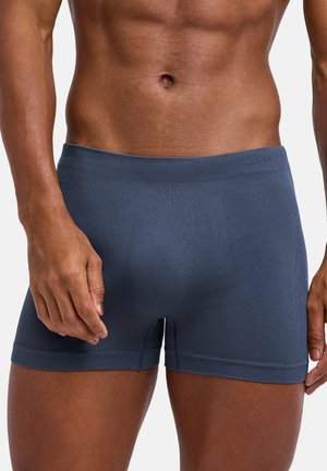 Mannelijke torso gekleed in strakzittende marineblauwe boxershorts, met zichtbare gespierde buik en beide handen langs het lichaam geplaatst.