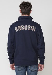 Sudadera azul marino con un cuello alto, que presenta el texto "KOROSHI" en letras estampadas y multicolores en la parte trasera. Mangas largas, ajuste casual.
