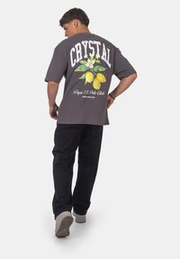 Šedé oversized tričko se žlutou potiskem citronu a květin na zadní straně, kombinované s černými kalhotami a světlými teniskami.