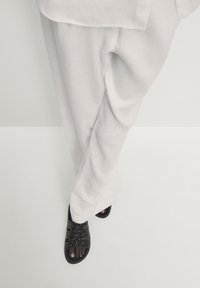 Sandales noires avec dessus texturé tressé, design à bout ouvert, associées à un pantalon blanc ample. Le fond est minimaliste et lumineux.