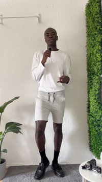 Pantalones cortos grises, camisa de manga larga blanca, zapatos negros, sosteniendo un micrófono, de pie contra una pared con vegetación, textura de tela gris claro.
