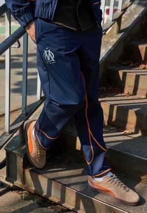Marineblauwe sportbroek met oranje stiksels, met een logo op de dij. Taupe sneakers met oranje accenten. Persoon die op een trap staat.