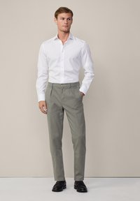 Weißes Hemd mit langen Ärmeln, kombiniert mit hellgrauen Hosen mit flacher Vorderseite und Hosentaschen. Schwarze Schuhe vervollständigen das Outfit.