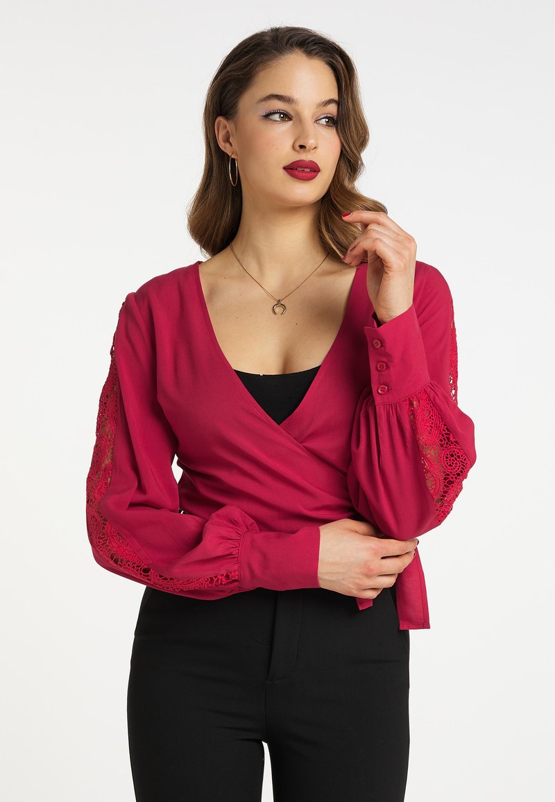 Fuchsia indpakningsbluse har lange puffærmer med blonde detaljer, en dyb V-udskæring, og knapper ved manchetterne, som er parret med sorte bukser.