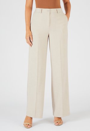 Pantalon large beige en tissu léger, avec fermeture à bouton, plis marqués et poches latérales, porté avec des chaussures à bouts ouverts.