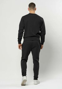 Muškarac stoji okrenut leđima, nosi crnu trenirku s bijelim tenisicama na jednostavnoj svijetloj pozadini.