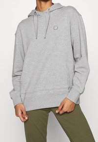 Sweatshirt à capuche gris en tissu doux, avec une poche avant et un petit logo circulaire sur la poitrine. Coupe décontractée avec des manches longues.