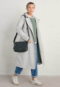 Lichtgrijze trenchcoat met knoopsluiting en brede revers, gecombineerd met een groene gebreide trui en blauwe spijkerbroek met opgerolde mouwen. Donkere tas en witte sneakers.