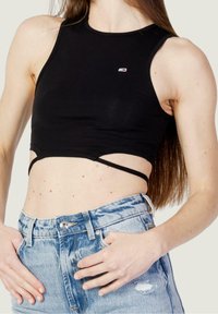 Kobieta w czarnej, krótkiej koszulce na ramiączkach i jasnoniebieskich, wystrzępionych dżinsach z wysokim stanem, z rękami w pobliżu kieszeni.