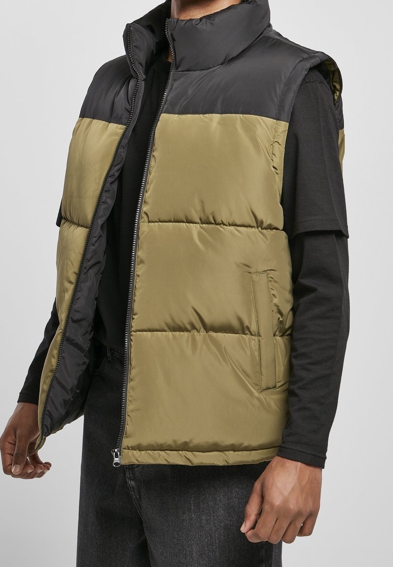 Vadderad utanärmad väst med en olivgrön kropp och svart övre del. Dragkedjestängning och sidofickor synliga. Slät, strukturerad tyg.