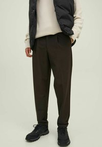 Pantalones oscuros de corte tapered en color marrón, con una textura suave, que cuentan con pliegues frontales y bolsillos laterales, combinados con zapatos de senderismo negros y un chaleco negro.