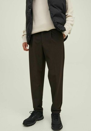 Pantalones oscuros de corte tapered en color marrón, con una textura suave, que cuentan con pliegues frontales y bolsillos laterales, combinados con zapatos de senderismo negros y un chaleco negro.