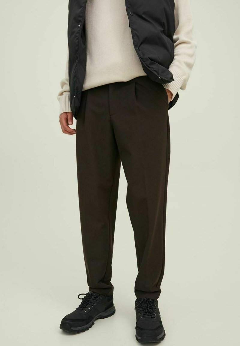 Pantalones oscuros de corte tapered en color marrón, con una textura suave, que cuentan con pliegues frontales y bolsillos laterales, combinados con zapatos de senderismo negros y un chaleco negro.