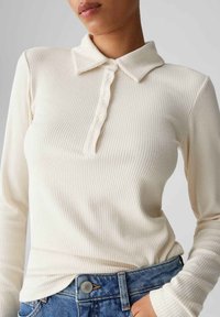 Top a maniche lunghe color beige chiaro con costine, colletto piegato e abbottonatura, abbinato a jeans. Tessuto strutturato e design aderente.