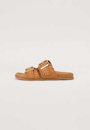 Tan slide sandals featuring two wide straps with cut-out patterns, gold-tone hardware, and a flat sole with textured details for grip.