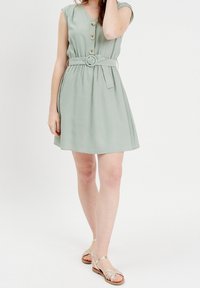 Robe courte vert menthe avec manches courtes à capuchon, boutonnée à l'avant, ceinture à la taille et jupe évasée. Portée avec des sandales métalliques à brides sur un fond uni.