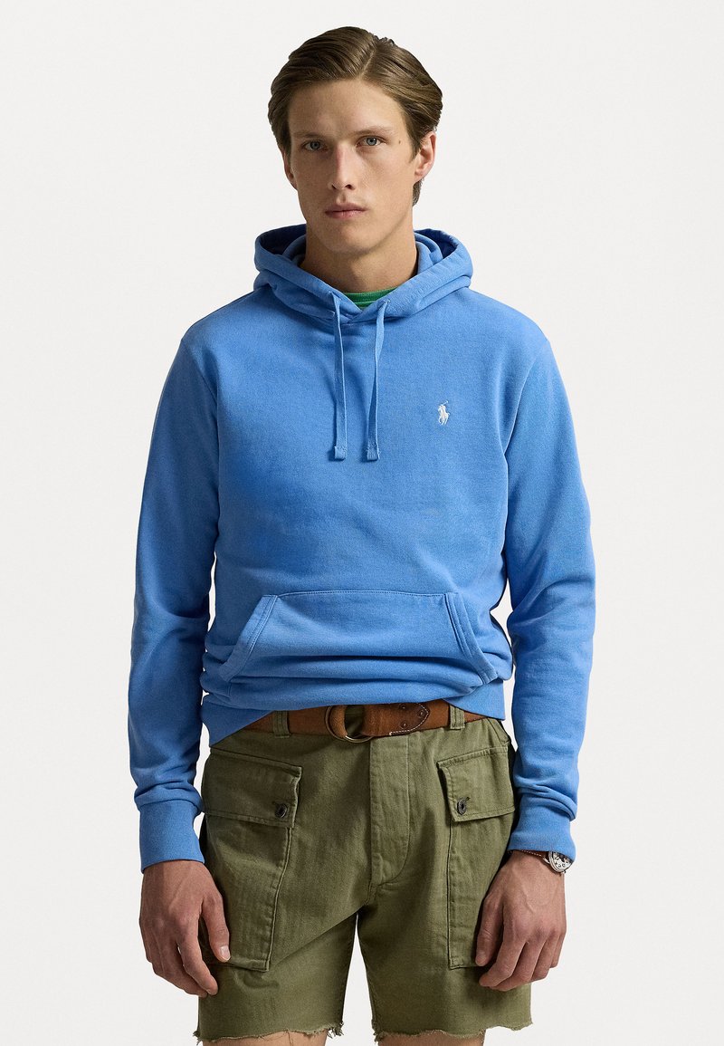 Blå bomuldshoodie med snoretræk, frontlomme og lille logo. Båret med olivengrønne cargo shorts med lommer med flap og frynset kant.