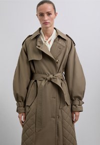 Gabardina marrón de gran tamaño con textura acolchada, cintura ceñida con cinturón, grandes botones y mangas amplias, fusionada sobre una blusa ligera.