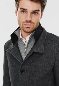 Czarny płaszcz z wełny w ciemnoszarym kolorze, z czarnym kołnierzem, zapinany na guziki, a pod nim szary sweter na zamek. Gładka tekstura, dopasowany krój.