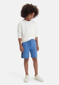 Παιδί με σγουρά μαλλιά που φοράει άσπρο μακρυμάνικο πουκάμισο, μπλε σορτς με πλευρικές ρίγες και άσπρα αθλητικά παπούτσια, στέκεται σε λευκό φόντο.