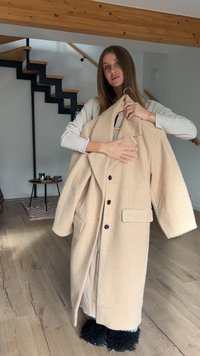 Beige, oversized, pluizige jas met een sjaalkraag, zwarte knopen en opgestikte zakken, gedragen door een persoon in een licht shirt. Zachte textuur zichtbaar.