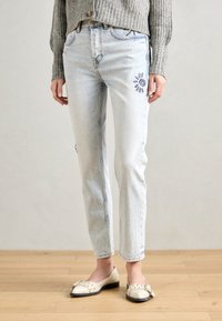 Lyseblå denimjeans med rett passform, prydet med broderte sol- og øyemønstre på låret, kombinert med kremfargede flate sko.