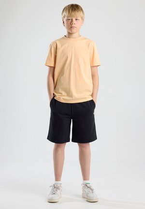 T-shirt en coton orange clair, shorts noirs, baskets blanches avec des accents gris. Design simple, coupe décontractée, poches sur les shorts.