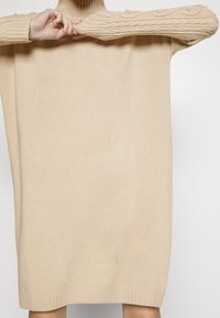 Personne portant une robe pull en maille beige oversize à manches longues et col roulé, ajustant la manche droite avec les deux mains.